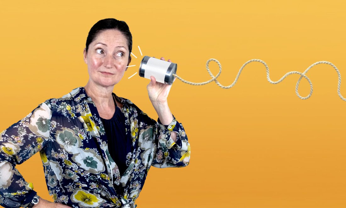 A woman holding a tin can attached to a piece of string to her ear in front of a yellow background